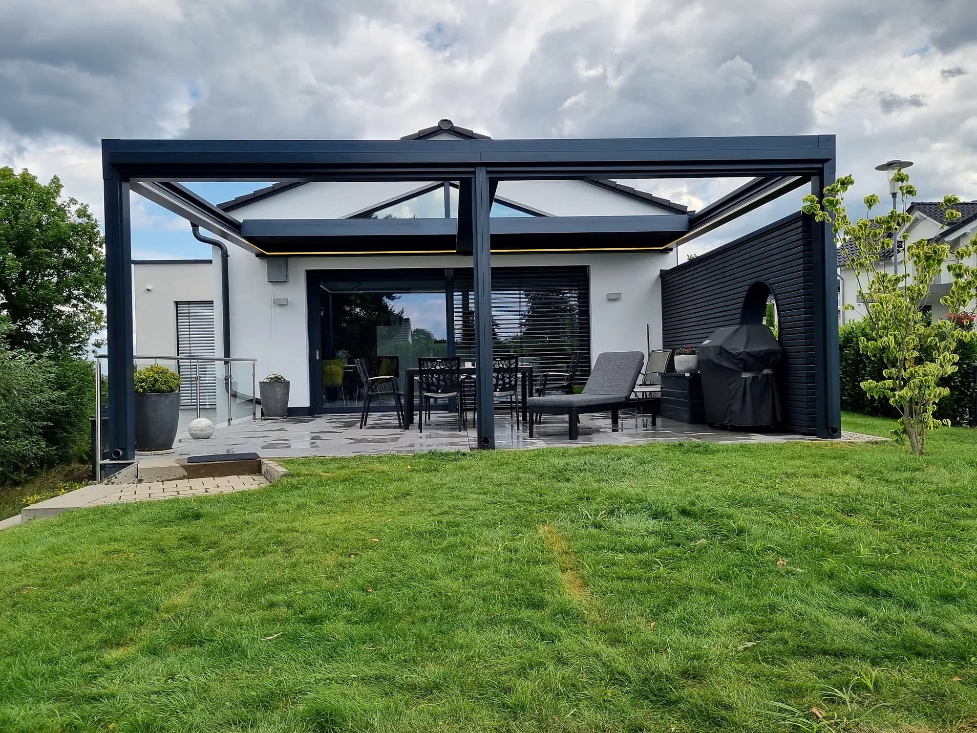 Pergola mit Glasdach und Senkrechtmarkisen auf einer Terrasse mit Rasen